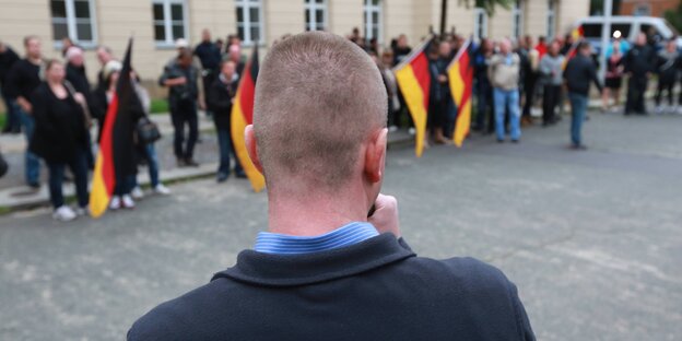 Ein Mann steht mit dem Rücken zur Kamera und spricht vor Menschen, die allesamt Deutschlandflaggen halten Ein Mann steht mit dem Rücken zur Kamera und spricht vor Menschen, die allesamt Deutschlandflaggen halten