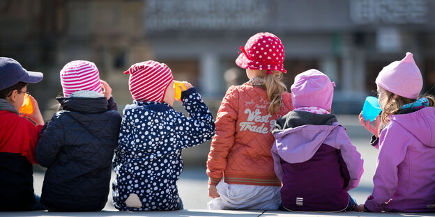 Ein paar Kinder sitzen in Jacken und Mützen gehüllt nebeneinander Ein paar Kinder sitzen in Jacken und Mützen gehüllt nebeneinander