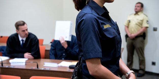 Der Angeklagt sitzt hinter einem Tisch und hält sich ein Papier vor das Gesicht. Im Vordergrund sieht man eine Justizbedienstete Der Angeklagt sitzt hinter einem Tisch und hält sich ein Papier vor das Gesicht. Im Vordergrund sieht man eine Justizbedienstete