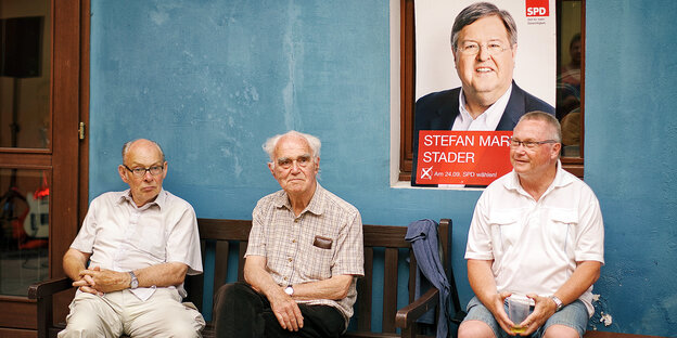drei alte Männer sitzen auf einer Bank vor einer Wand, darauf ein SPD-Plakat mit dem Gesicht von Klaus Maria Stader drei alte Männer sitzen auf einer Bank vor einer Wand, darauf ein SPD-Plakat mit dem Gesicht von Klaus Maria Stader