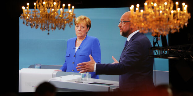 Auf einer Leinwand sind Merkel und Schulz zu sehen, vor ihnen hängen Kronleuchter Auf einer Leinwand sind Merkel und Schulz zu sehen, vor ihnen hängen Kronleuchter