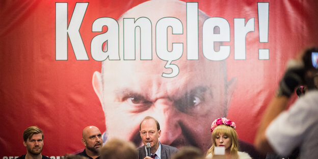 Auf einem roten Plakat steht „Kancler!“, davor sitzen Menschen Auf einem roten Plakat steht „Kancler!“, davor sitzen Menschen