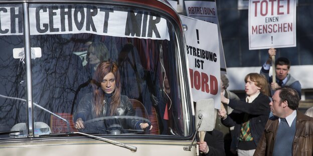 Eine rothaarige Frau lenkt einen Bus mit einem Banner, auf dem "Mein Bauch gehört mir" steht, um sie herum demonstrieren Abtreibungsgegner mit Plakaten Eine rothaarige Frau lenkt einen Bus mit einem Banner, auf dem "Mein Bauch gehört mir" steht, um sie herum demonstrieren Abtreibungsgegner mit Plakaten