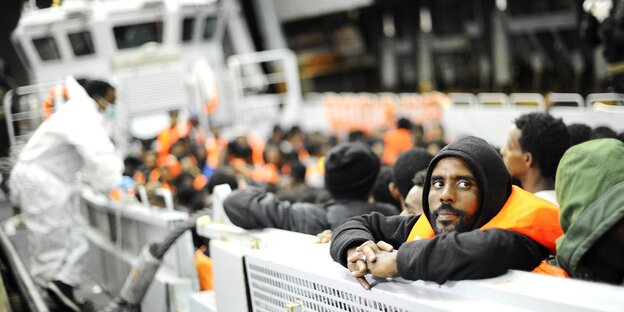 Flüchtlinge an Deck eines weißen Schiffes, einer hat die Arme auf der Reling verschränkt und blickt den Betrachter an Flüchtlinge an Deck eines weißen Schiffes, einer hat die Arme auf der Reling verschränkt und blickt den Betrachter an