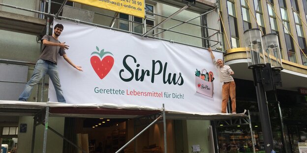 Zwei Initiatoren des Supermakrtes präsentieren ein Plakat, das über dem Laden hängt.