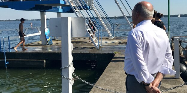 Ein Mann im Hemd schaut auf einen Sprungturm vor einem See Ein Mann im Hemd schaut auf einen Sprungturm vor einem See