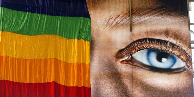 Ein Plakat mit einem großen Gesicht neben einer Regenbogenfahne Ein Plakat mit einem großen Gesicht neben einer Regenbogenfahne