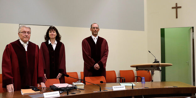 Drei Menschen in Robe stehen hinter einem Tisch in einem Gerichtsaal - oben rechts hängt ein Kreuz an der Wand Drei Menschen in Robe stehen hinter einem Tisch in einem Gerichtsaal - oben rechts hängt ein Kreuz an der Wand