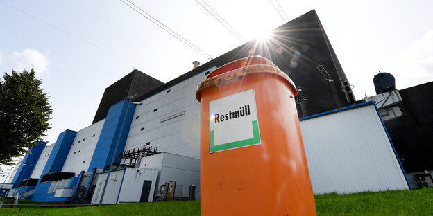 Vor einem Atomkraftwerk steht eine orangefarbene Mülltonne, auf der „Restmüll“ steht Vor einem Atomkraftwerk steht eine orangefarbene Mülltonne, auf der „Restmüll“ steht