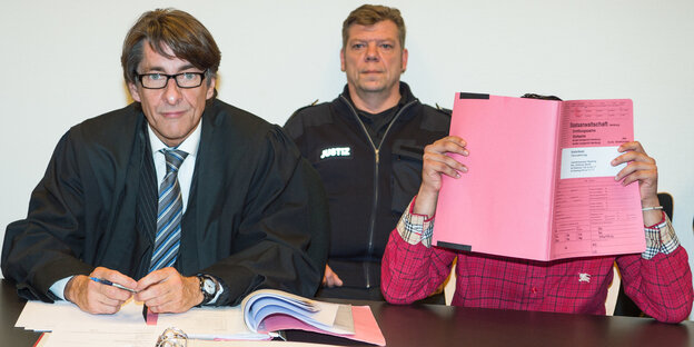 Ein Anwalt und jemand, der sein Gesicht mit einem Heft verdeckt sitzen an einem Tisch, im Hintergrund sitzt ein Polizist Ein Anwalt und jemand, der sein Gesicht mit einem Heft verdeckt sitzen an einem Tisch, im Hintergrund sitzt ein Polizist