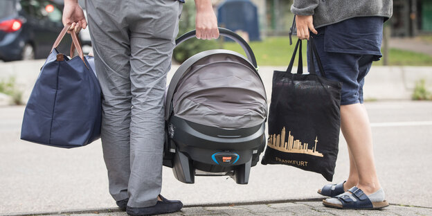 Ein Mann und eine Frau sind von hinten und von der Hüfte abwärts zu sehen, sie tragen jeder eine schwere Tasche und er eine Kindertrage Ein Mann und eine Frau sind von hinten und von der Hüfte abwärts zu sehen, sie tragen jeder eine schwere Tasche und er eine Kindertrage