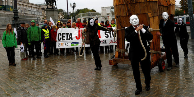 Protest gegen Ceta, Menschen mit Bannern und Menschen mit Masken, die ein Holzpferd ziehen Protest gegen Ceta, Menschen mit Bannern und Menschen mit Masken, die ein Holzpferd ziehen