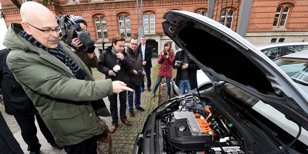 Mann zeigt auf offenen Motorraum. Darin ist ein Elektromotor Mann zeigt auf offenen Motorraum. Darin ist ein Elektromotor