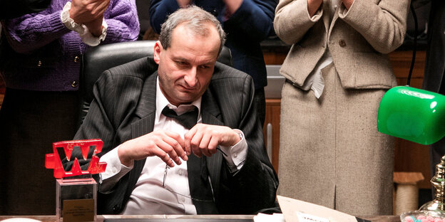 Ein Mann sitzt an einem Schreibtisch, hinter ihm stehen Menschen Ein Mann sitzt an einem Schreibtisch, hinter ihm stehen Menschen