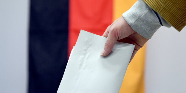 Jemand steckt einen Wahlzettel in eine Box, dahinter sieht man eine Deutschland-Flagge Jemand steckt einen Wahlzettel in eine Box, dahinter sieht man eine Deutschland-Flagge