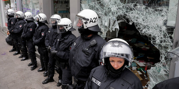 Polizisten in Schutzkleidung und mit Helmen stehen vor einer zerstörten Glasscheibe in einer Reihe nebeneinander Polizisten in Schutzkleidung und mit Helmen stehen vor einer zerstörten Glasscheibe in einer Reihe nebeneinander