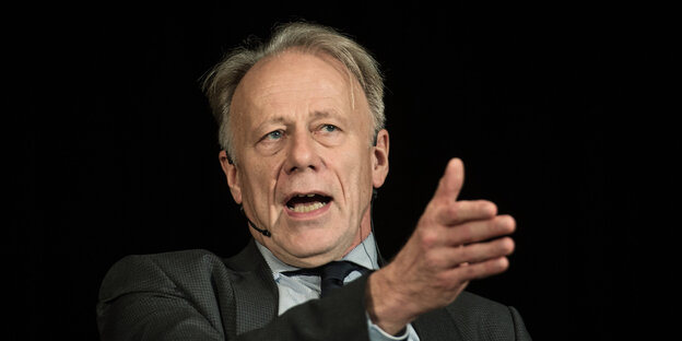 Jürgen Trittin sitzt vor schwarzem Hintergrund und weist mit dem rechten Arm nach links Jürgen Trittin sitzt vor schwarzem Hintergrund und weist mit dem rechten Arm nach links