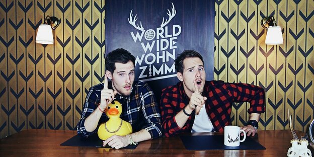 Die beiden Moderatoren von "World Wide Wohnzimmer" in ihrem Youtubestudio an einem Tisch mit bunter Tapete dahinter Die beiden Moderatoren von "World Wide Wohnzimmer" in ihrem Youtubestudio an einem Tisch mit bunter Tapete dahinter