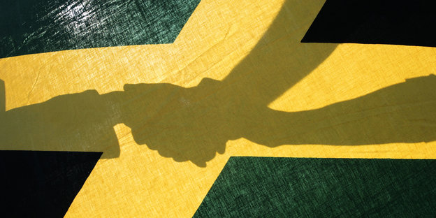Die Schatten von drei Händen, die einander halten. Davor eine Jamaika-Flagge. Die Schatten von drei Händen, die einander halten. Davor eine Jamaika-Flagge.