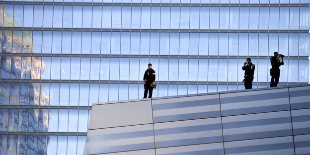 Drei Polizeibeamte stehen auf einem spiegelnden Haus, im Hintergrund ein höheres, ebenfalls spiegelndes Haus Drei Polizeibeamte stehen auf einem spiegelnden Haus, im Hintergrund ein höheres, ebenfalls spiegelndes Haus