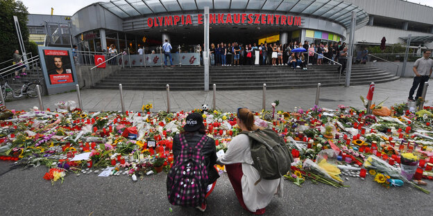 Zwei Frauen kniene vor den niedergelegten Blumen vor dem Olympiaeinkaufszentrum