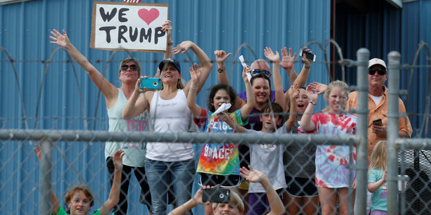 Menschen winken hinter einem Stacheldrahtzaun und halten ein "We love Trump"-Schild in die Höhe Menschen winken hinter einem Stacheldrahtzaun und halten ein "We love Trump"-Schild in die Höhe