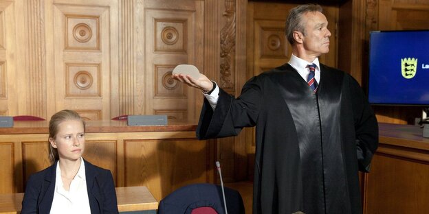 Der Anwalt hält im Gerichtssaal mit theatralischer Geste ein Implantat hoch. Die Klägerin sitzt daneben Der Anwalt hält im Gerichtssaal mit theatralischer Geste ein Implantat hoch. Die Klägerin sitzt daneben