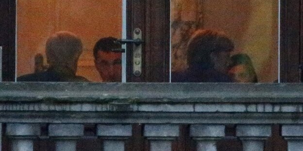 Vier Menschen sitzen hinter einer dicken Balkonbrüstung hinter einer Fensterscheibe