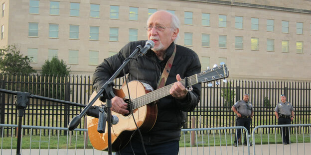 Peter Yarrow steht vor dem Pentagon, spielt Guitarre und singt in ein Mikrofon Peter Yarrow steht vor dem Pentagon, spielt Guitarre und singt in ein Mikrofon