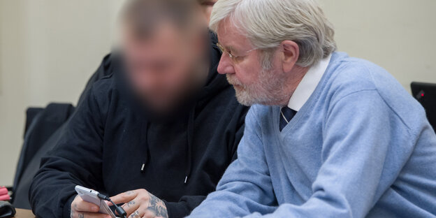 Ein Mann mit grauen Haaren und blauem Pullover neben einem dunkel gekleideten Mann, dessen Gesicht unkenntlich gemacht wurde Ein Mann mit grauen Haaren und blauem Pullover neben einem dunkel gekleideten Mann, dessen Gesicht unkenntlich gemacht wurde