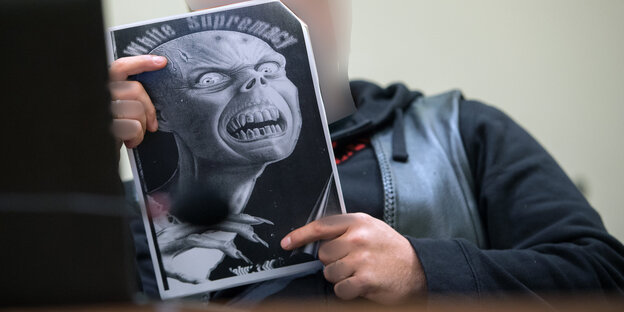 Eine white-Supremacy-Monstermaske ist auf einem Poster abgedruckt, das ein Mann in einem Gerichtssaal in den Händen hält Eine white-Supremacy-Monstermaske ist auf einem Poster abgedruckt, das ein Mann in einem Gerichtssaal in den Händen hält