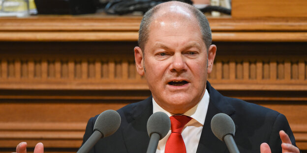 Ein Mann mit Halbglatze und roter Krawatte steht gestikulierend an einem Mikrofon Ein Mann mit Halbglatze und roter Krawatte steht gestikulierend an einem Mikrofon