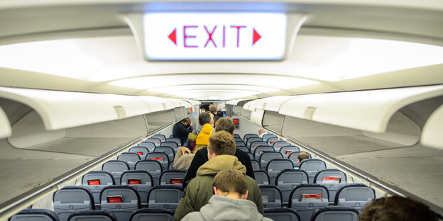 Menschen zwischen Sitzreihen in einem Flugzeug unter einem Schild, auf dem „Exit“ steht