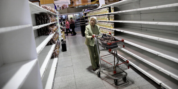 Eine ältere Frau schiebt einen Einkaufswagen durch leere Supermarktregale Eine ältere Frau schiebt einen Einkaufswagen durch leere Supermarktregale