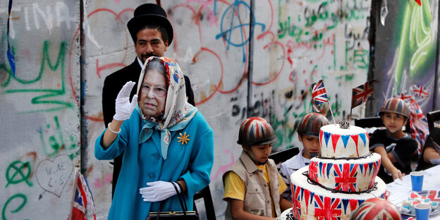Eine Frau als Queen Elizabeth II verkleidet, geht winkend an einer Mauer vorbei Eine Frau als Queen Elizabeth II verkleidet, geht winkend an einer Mauer vorbei