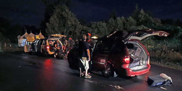 Autos mit Kofferräumen voller Gepäck stehen nachts auf einer dunklen Straße, die Fahrer stehen daneben Autos mit Kofferräumen voller Gepäck stehen nachts auf einer dunklen Straße, die Fahrer stehen daneben