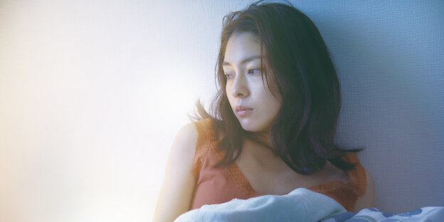 Eine Frau sitzt im Bett und lehnt an der Wand, die Bettdecke brusthoch gezogen, mit melancholischem Blick zur Seite Eine Frau sitzt im Bett und lehnt an der Wand, die Bettdecke brusthoch gezogen, mit melancholischem Blick zur Seite