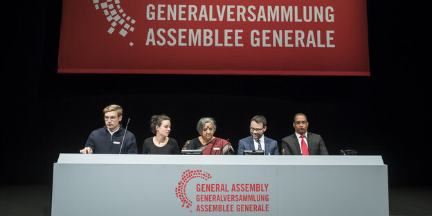 Fünf Menschen sitzen an einem Tisch, auf dem "Generalversammlung" steht Fünf Menschen sitzen an einem Tisch, auf dem "Generalversammlung" steht