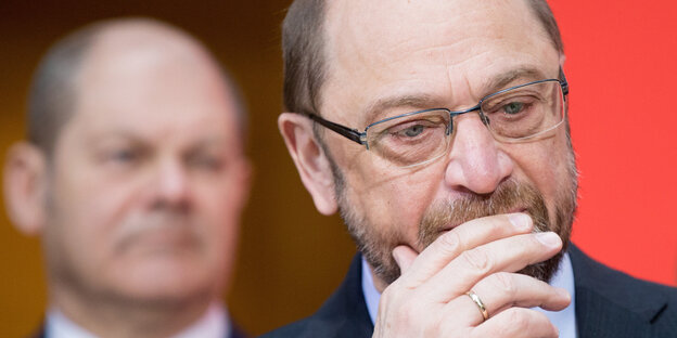 Ein Mann mit Brille guckt nachdenklich, im Hintergrund sieht man einen weiteren Mann Ein Mann mit Brille guckt nachdenklich, im Hintergrund sieht man einen weiteren Mann