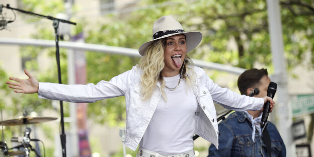 Miley cirus steht auf einer Bühne, breitet ihre Arme aus und steckt die Zunge heraus Miley cirus steht auf einer Bühne, breitet ihre Arme aus und steckt die Zunge heraus