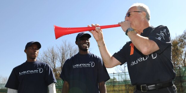 Franz Beckenbauer bläst in eine rote Vuvuzela. Zwei Männer beobachten ihn dabei Franz Beckenbauer bläst in eine rote Vuvuzela. Zwei Männer beobachten ihn dabei