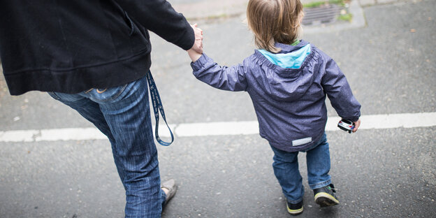 Ein Erwachsener hält ein Kind an der Hand, sie laufen eine Straße entlang Ein Erwachsener hält ein Kind an der Hand, sie laufen eine Straße entlang
