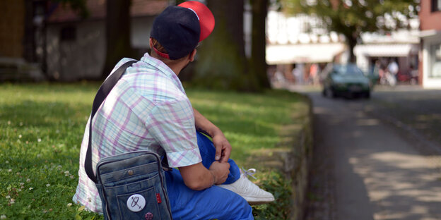Ein Mensch sitzt auf dem Rasen am Rand einer Straße, sein Gesicht ist abgewandt Ein Mensch sitzt auf dem Rasen am Rand einer Straße, sein Gesicht ist abgewandt