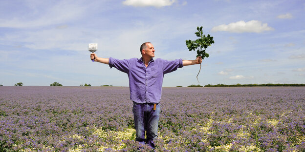 Bill Drummond steht in einem Feld und hält einen Zweig in der einen, einen Pinsel in der anderen Hand Bill Drummond steht in einem Feld und hält einen Zweig in der einen, einen Pinsel in der anderen Hand