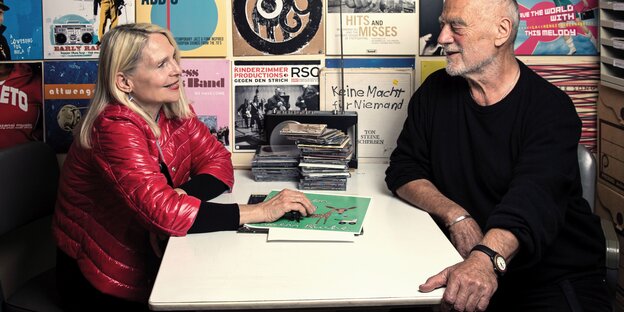 ein Mann und eine Frau sitzen sich an einem Tisch gegenüber, hinter ihnen eine Wand voller LP-Cover ein Mann und eine Frau sitzen sich an einem Tisch gegenüber, hinter ihnen eine Wand voller LP-Cover