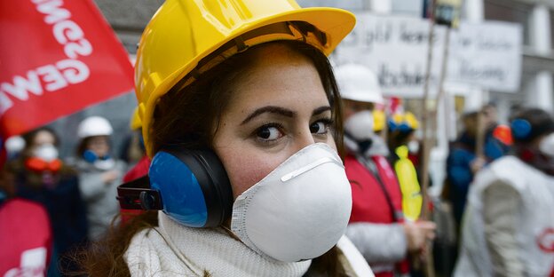 Eine Frau mit Helm, Atemschutz und Kopfhörern protestiert gegen den schlechten Zustand einer Berliner Schule Eine Frau mit Helm, Atemschutz und Kopfhörern protestiert gegen den schlechten Zustand einer Berliner Schule