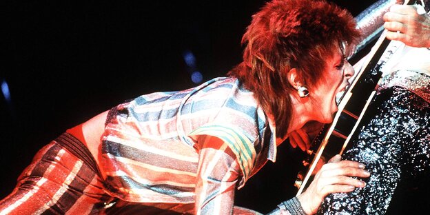 David Bowie mit Rothaar-Frisur beißt in eine Gitarre David Bowie mit Rothaar-Frisur beißt in eine Gitarre