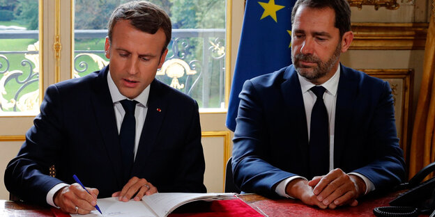 Emmanuel Macron und Christophe Castaner sitzen nebeneinander an eine Tisch. Macron unterschreibt ein Papier Emmanuel Macron und Christophe Castaner sitzen nebeneinander an eine Tisch. Macron unterschreibt ein Papier