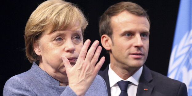 Angela Merkel bläst die Backen auf und hebt die Hand. Emmanuel Macron steht im Hintergrund Angela Merkel bläst die Backen auf und hebt die Hand. Emmanuel Macron steht im Hintergrund