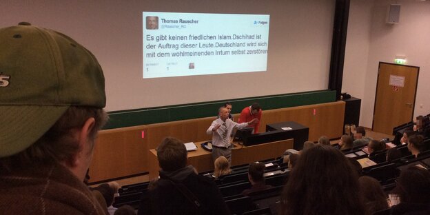 Ein Mann mit erhobenen Armen steht in einem Vorlesungssaal Ein Mann mit erhobenen Armen steht in einem Vorlesungssaal